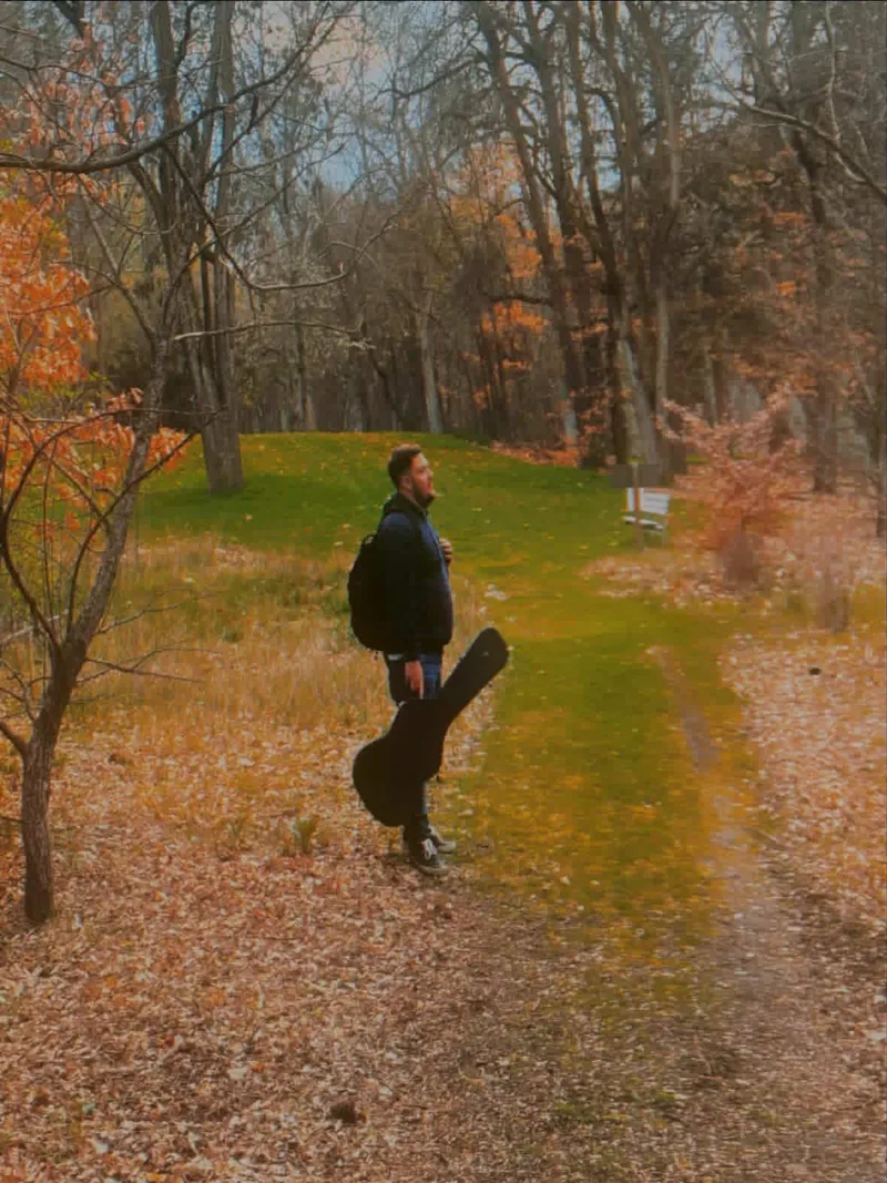 Dustin Leonard Jolley walking with guitar case through autumn leaves